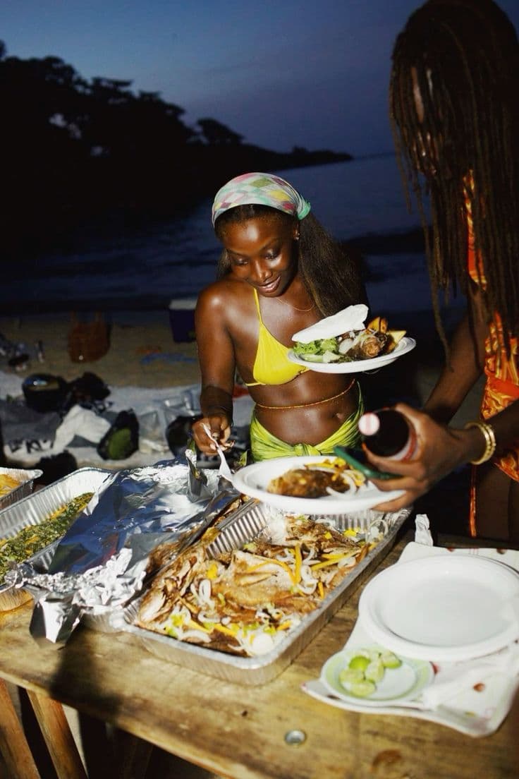 Two women serve grilled fish and sides from large trays at a nighttime beach gathering.